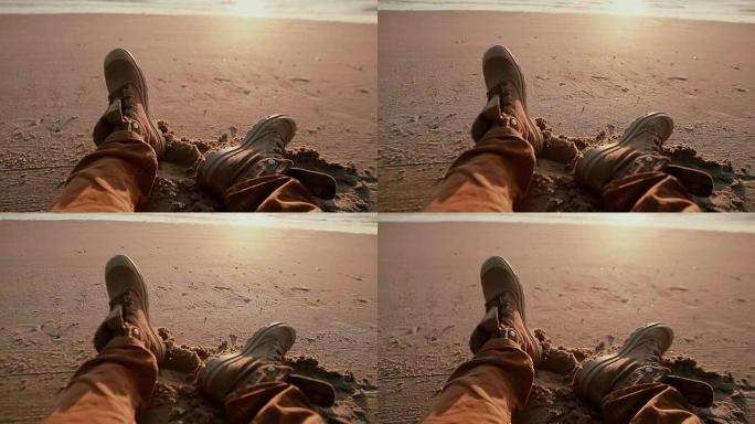 两只破旧的靴子的特写镜头，属于一个男人在沙滩上休息，看着美丽的日落