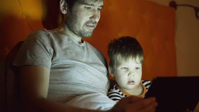 年轻的父亲和他的小儿子晚上在家躺在床上用平板电脑看卡通电影
