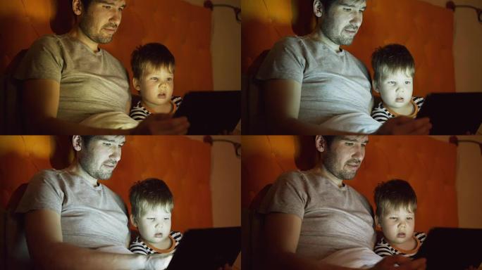 年轻的父亲和他的小儿子晚上在家躺在床上用平板电脑看卡通电影
