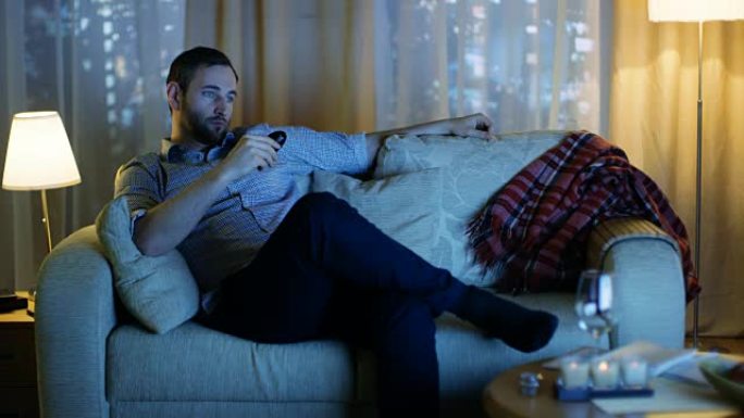 晚上，中年男子坐在沙发上看电视，用遥控器换频道。在具有摩天大楼的大城市的背景窗口视图中。
