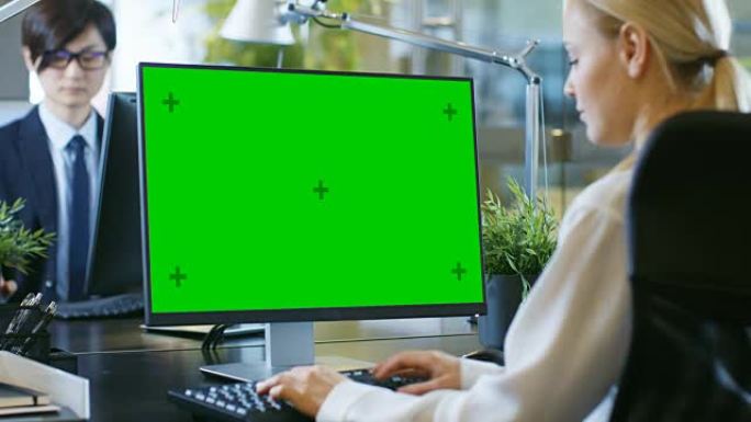 在办公室里，女商人在办公桌前工作，使用带有绿屏模型的个人计算机。同事坐在她对面。现代外观的房间，玻璃