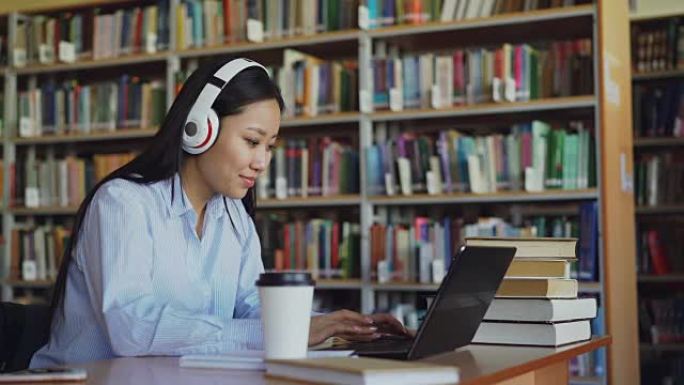 美丽的亚洲女学生戴着耳机听音乐，坐在图书馆的办公桌上，在学校项目中工作。年轻女孩在笔记本电脑上打字，