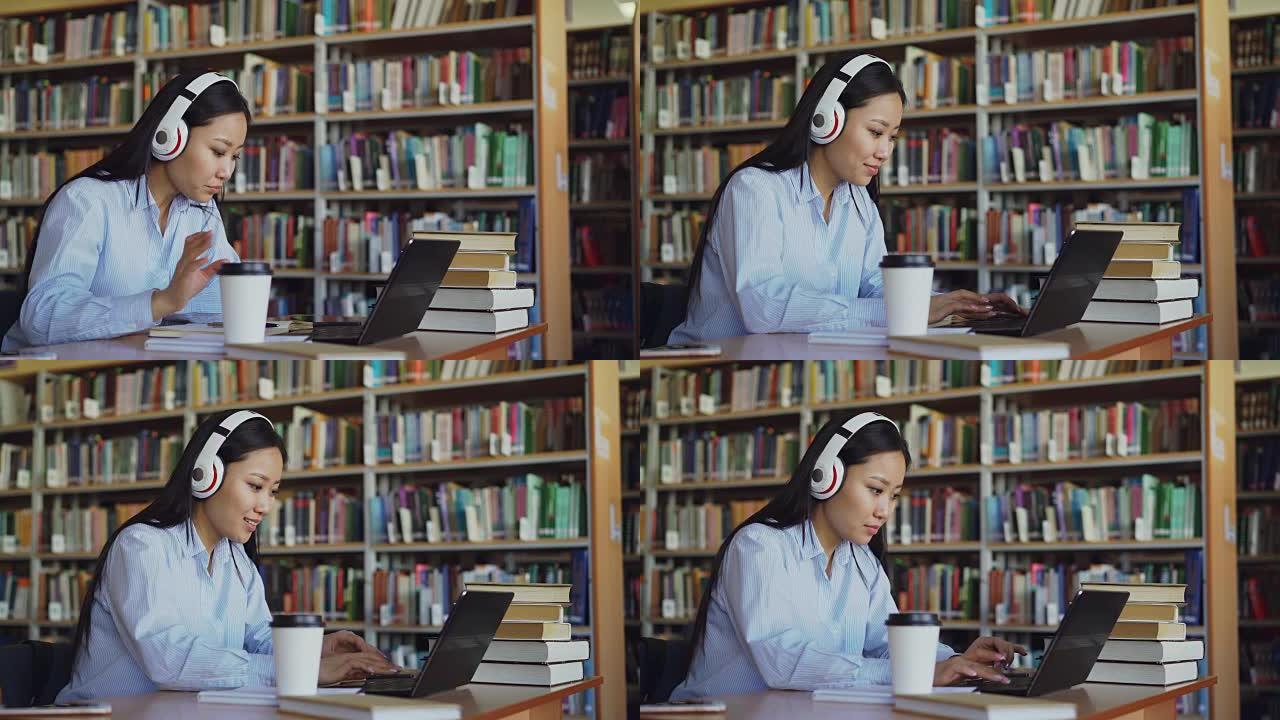 美丽的亚洲女学生戴着耳机听音乐，坐在图书馆的办公桌上，在学校项目中工作。年轻女孩在笔记本电脑上打字，