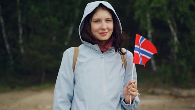 迷人的挪威体育迷的慢动作肖像挥舞着挪威的官方旗帜，站在森林里微笑着。人、民族和国家的概念。