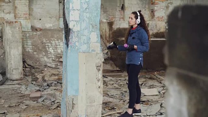 苗条女孩现代画家的俯仰镜头用气雾剂涂料在废墟仓库的高柱上画涂鸦，用耳机听音乐。