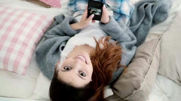 年轻漂亮的女人躺在床上，用智能手机看照片。女孩看着镜头微笑