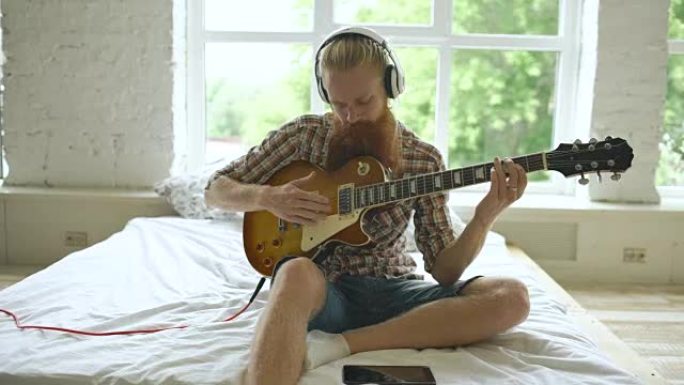 迷人的大胡子男人坐在床上学习用平板电脑在家里的现代卧室弹吉他