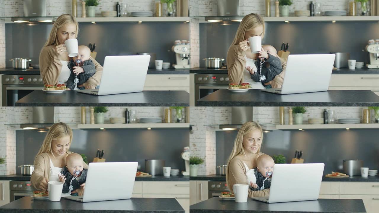微笑的母亲坐在笔记本电脑前的厨房桌子上，将好奇的孩子抱在膝盖上。母亲也从杯子里喝了一杯。
