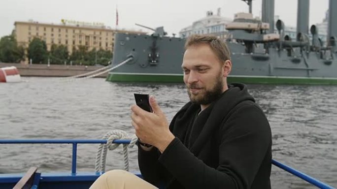 快乐英俊的男人在圣彼得堡观光。在巡洋舰Aurora面前自拍，使用他的智能手机slow mo