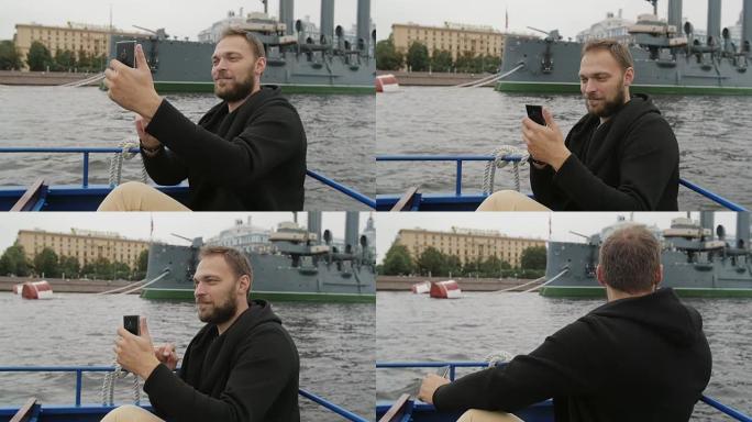 快乐英俊的男人在圣彼得堡观光。在巡洋舰Aurora面前自拍，使用他的智能手机slow mo