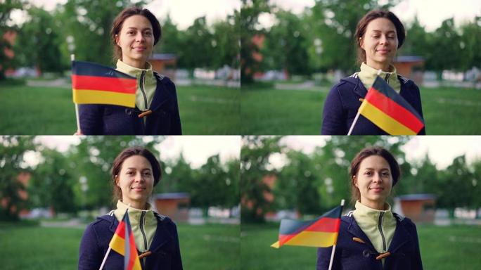 快乐的年轻女子的慢动作肖像挥舞着德国官方国旗，看着相机，站在美丽的绿色公园，树木和草坪。