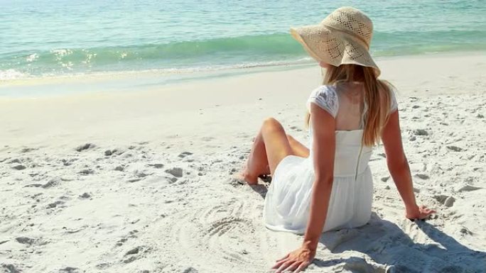 金发女子坐在沙滩上看着大海的后视图