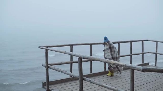 穿着胶靴和格子的小女孩站在海边的木墩上的背景图。雾蒙蒙的大风天独自一人的孩子