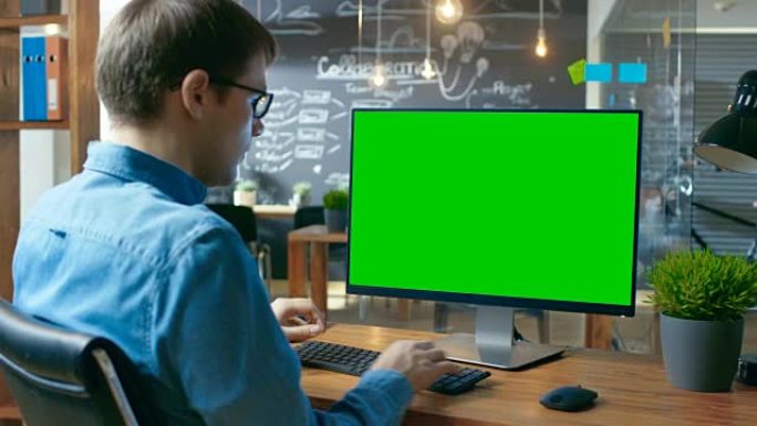 年轻人在带有模拟绿屏的个人计算机上的办公桌上工作。在后台，他的同事在创意办公室工作。