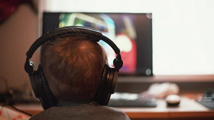 坐在椅子上的小男孩戴着大耳机仔细地看着电脑显示器上的东西