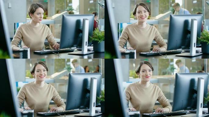 美丽年轻的白人女人坐在她的桌子和工作在一个个人电脑。在后台与同事忙碌的办公室。