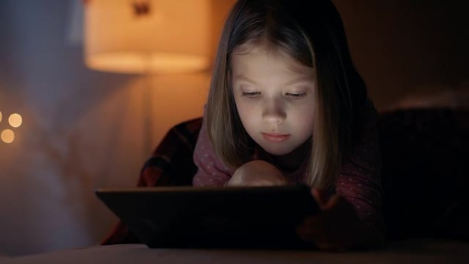 可爱的小女孩晚上躺在床上，她拿着平板电脑。她的夜灯打开了。