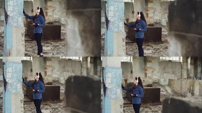 现代城市画家的倾斜镜头在废弃仓库的高柱上用气溶胶涂料绘制涂鸦，并用耳机听音乐。