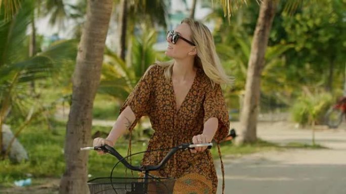 穿着连衣裙的美女骑着自行车在路上穿过正宗的小镇。南亚自然，棕榈树生长在路边。特写慢动作。