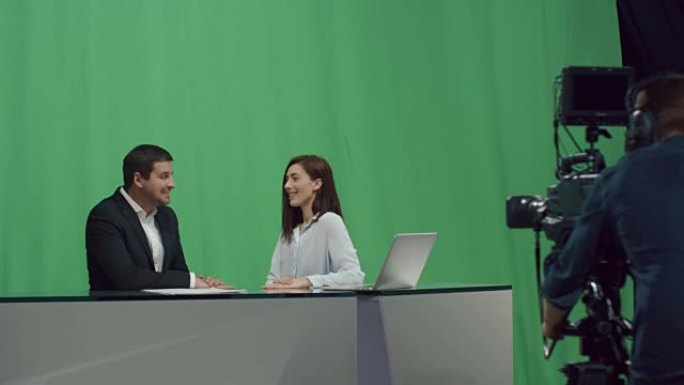 男性和女性广播公司的后台镜头，他们坐在桌子旁，在后台的模拟绿屏上聊天。