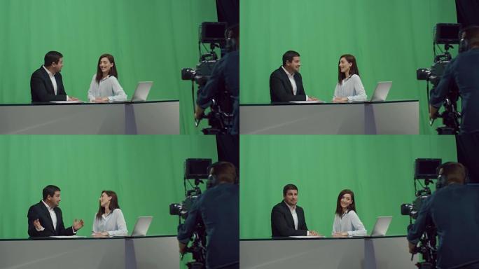 男性和女性广播公司的后台镜头，他们坐在桌子旁，在后台的模拟绿屏上聊天。