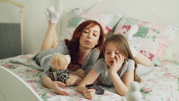 快乐快乐的母亲，可爱的女儿在电视上看卡通电影，早上躺在家里的床上，在舒适的卧室里，用遥控器微笑着