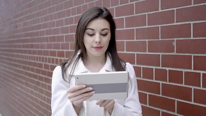 年轻漂亮的棕发女人在砖墙前用平板电脑工作。漂亮的白人女人在白色外套。