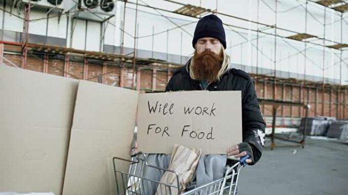 年轻的无家可归者的肖像，用纸板看着相机，想为站在购物车附近的食物工作
