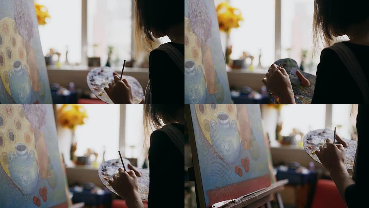 女艺术家的特写镜头将画笔与画笔混合在调色板中，然后在艺术工作室的画布上画静物画