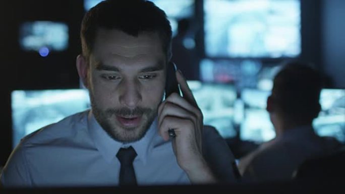 安全员在一个充满显示屏的黑暗监控室里的计算机上工作时正在打电话。