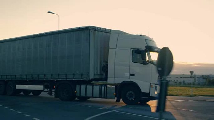 带有货运拖车的半卡车在高速公路上行驶的后续镜头。白色卡车在清晨驶过工业仓库区。