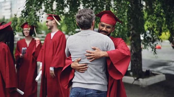 骄傲的老师正在祝贺学生，在毕业典礼那天穿着传统的研究生服装站在校园里握手并拥抱他。教育和人的概念。