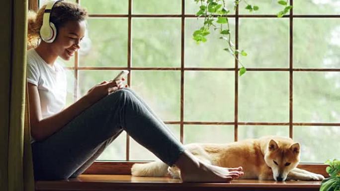 迷人的年轻女子正在通过无线耳机听音乐，并在智能手机上选择歌曲，她的宠物狗躺在主人附近的窗台上。