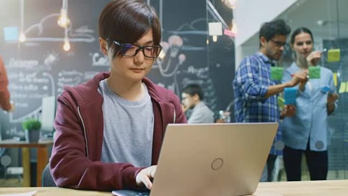 有才华的亚洲开发人员坐在笔记本电脑上工作。在繁忙的办公室中，有创造力的人在个人计算机上工作，在带有粘