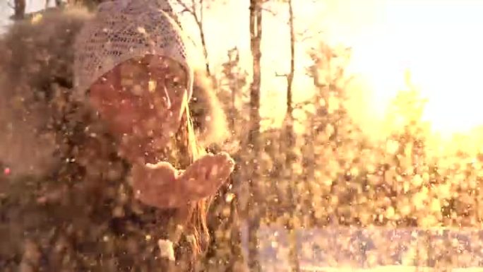 慢动作: 欢快的女孩在下雪的冬天日落时将雪花吹入相机