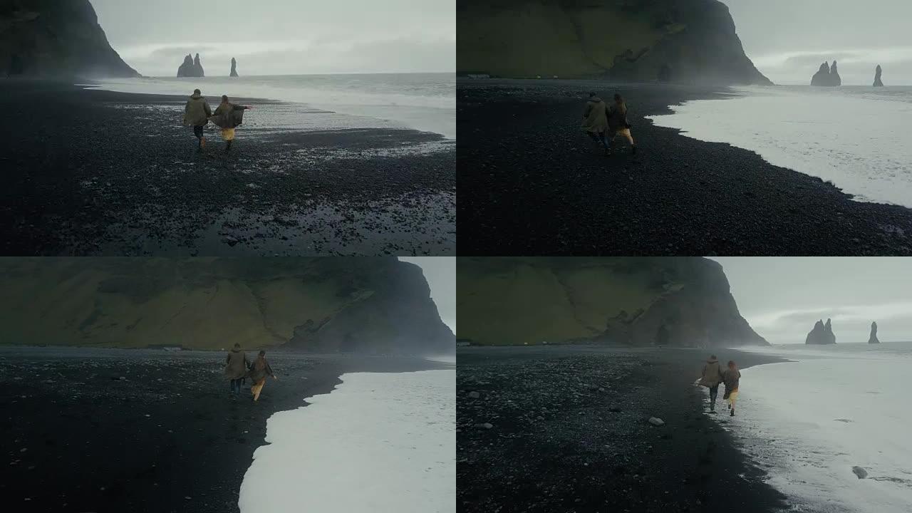 在冰岛巨魔脚趾附近的黑色火山海滩上奔跑的年轻时髦夫妇的后鸟瞰图