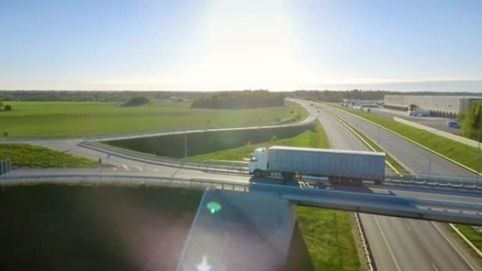 白色半卡车的鸟瞰图，货物拖车通过高速公路立交桥/桥梁。十八惠勒是新的，在后台可以看到装载仓库。