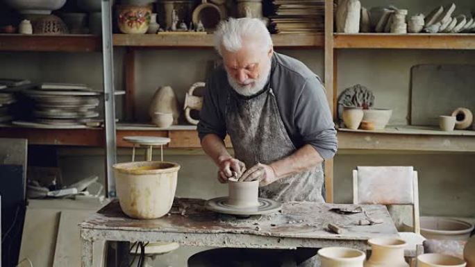 高级专业陶工正在用旋转的陶工轮上的粘土制作碗，使湿的粘土器皿变宽并变平。背景不同的手工陶瓷人物。