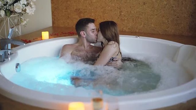 穿着泳衣的年轻有魅力的人在日间水疗中心用泡沫在热水浴缸中拥抱和亲吻。浪漫关系、激情和健康概念。