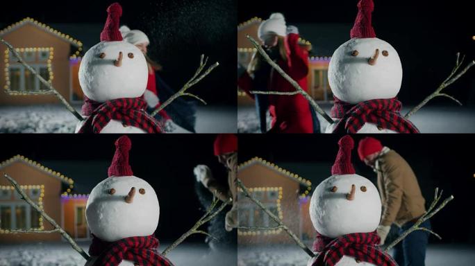 可爱的雪人站在房子的后院。在背景中，恋爱中的情侣在雪球中玩耍。冬天晚上的乐趣。