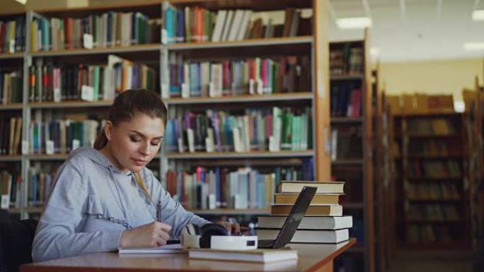 年轻美丽的微笑学生女孩坐在宽敞的大图书馆的办公桌前，那里堆满了书。她在抄写本上写东西，笔记本电脑就在