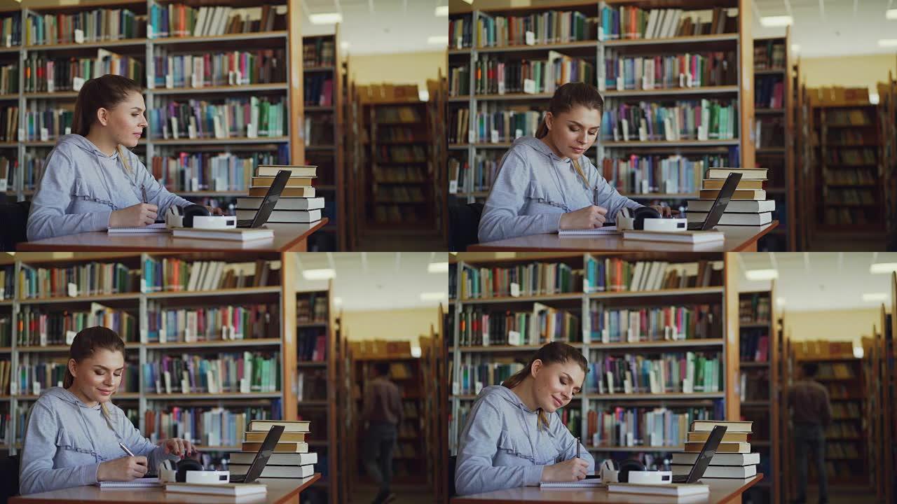 年轻美丽的微笑学生女孩坐在宽敞的大图书馆的办公桌前，那里堆满了书。她在抄写本上写东西，笔记本电脑就在