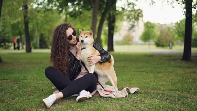 快乐女孩骄傲的狗主人爱抚和亲吻她的宠物坐在公园的草地上，而动物正在享受爱和关怀。绿树和草坪是可见的。