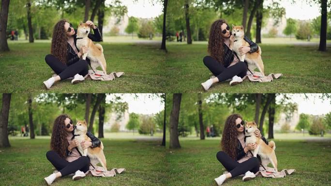 快乐女孩骄傲的狗主人爱抚和亲吻她的宠物坐在公园的草地上，而动物正在享受爱和关怀。绿树和草坪是可见的。