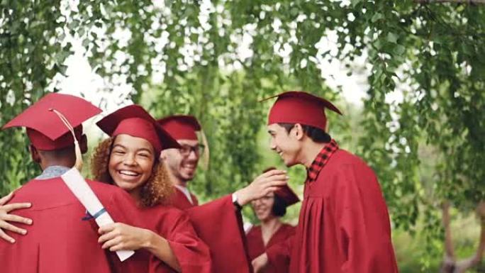 毕业典礼结束后，快乐的毕业学生多种族团体拥抱并击掌，穿着灰泥板和长袍的女孩和男孩都在笑着玩。