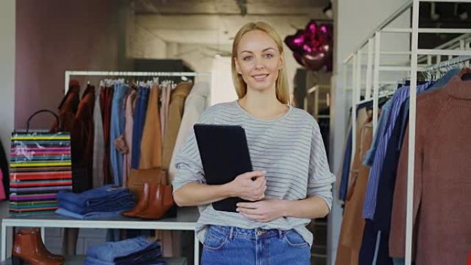 成功的年轻女商人的肖像站在她的服装店，拿着平板电脑，微笑着看着相机。宽敞的精品店，背景是女装。