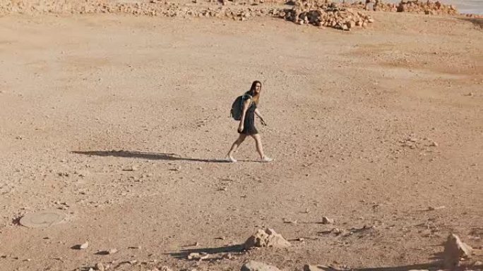背着背包的女人走向沙漠废墟。年轻的高加索女性旅行者在干燥的沙子、岩石、墙壁上。马萨达以色列4K