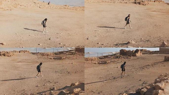背着背包的女人走向沙漠废墟。年轻的高加索女性旅行者在干燥的沙子、岩石、墙壁上。马萨达以色列4K