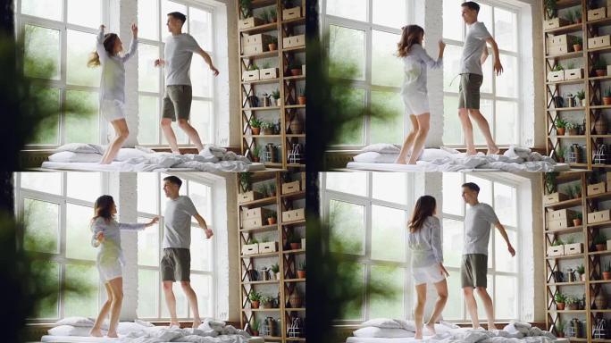 年轻的已婚夫妇在大窗户的轻便房间里的双人床上跳跃和跳舞的慢动作，快乐的人们在开心和大笑。