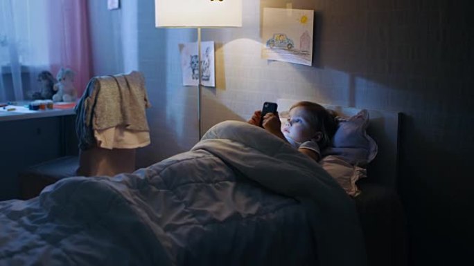 可爱的小女孩躺在床上，用智能手机看卡通。她的落地灯打开了。